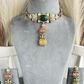 Gold-toned traditional Indian jewelry set featuring a choker necklace and matching earrings, displayed on a gray velvet mannequin bust. The choker has a large green oval stone, a central coral-pink round stone, a yellow square stone, and small white pearl-like accents.