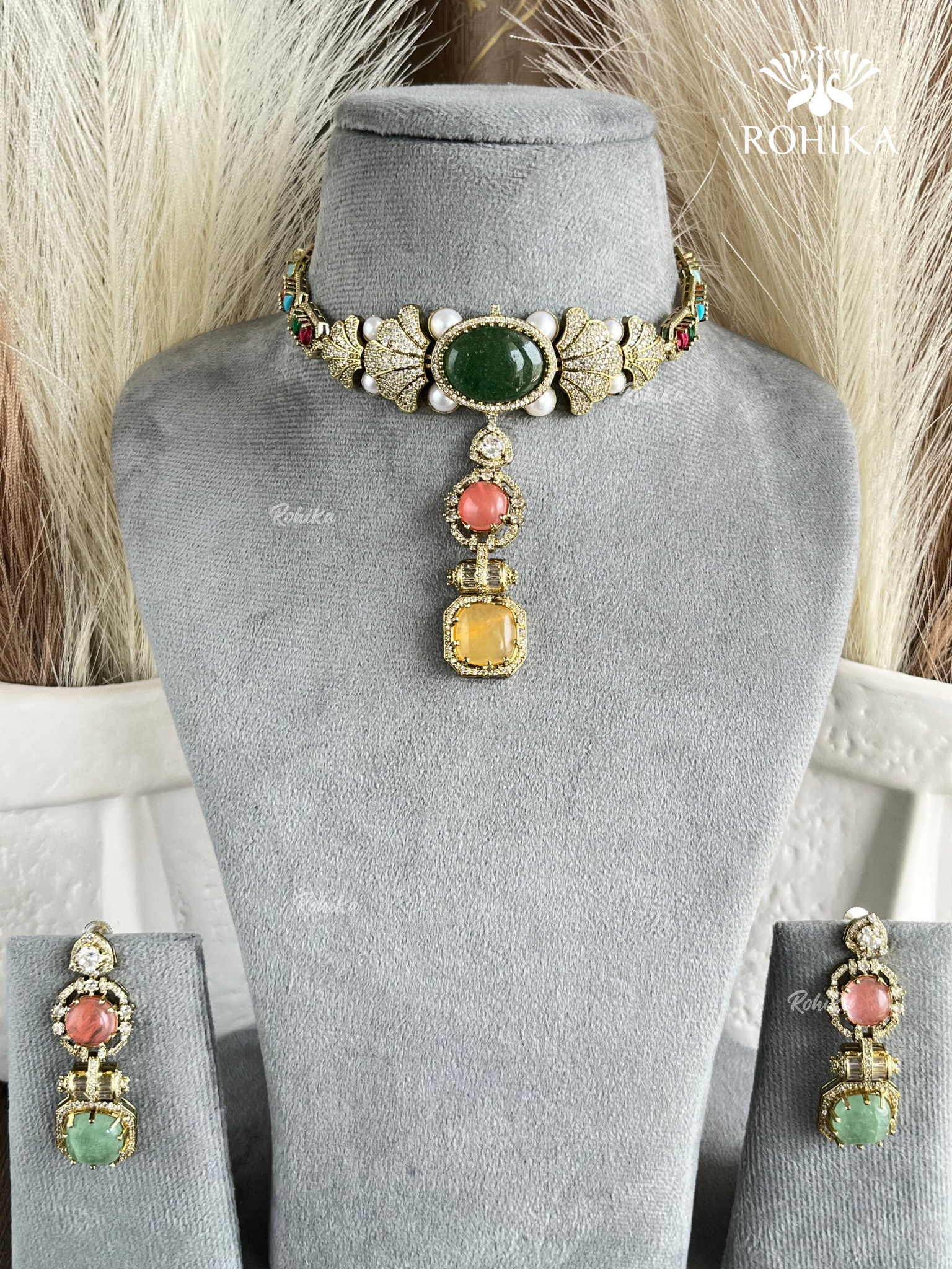 Gold-toned traditional Indian jewelry set featuring a choker necklace and matching earrings, displayed on a gray velvet mannequin bust. The choker has a large green oval stone, a central coral-pink round stone, a yellow square stone, and small white pearl-like accents.