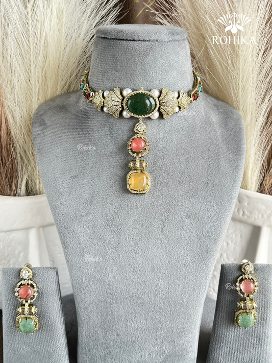 Gold-toned traditional Indian jewelry set featuring a choker necklace and matching earrings, displayed on a gray velvet mannequin bust. The choker has a large green oval stone, a central coral-pink round stone, a yellow square stone, and small white pearl-like accents.