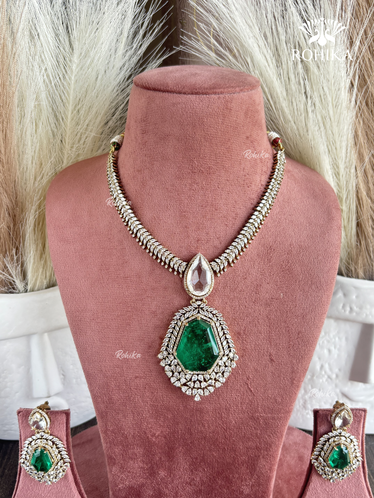 A gold-toned traditional Indian jewelry set displayed on a dusty rose velvet mannequin bust, featuring a necklace with a large green teardrop pendant, matching drop earrings, and a necklace stand.