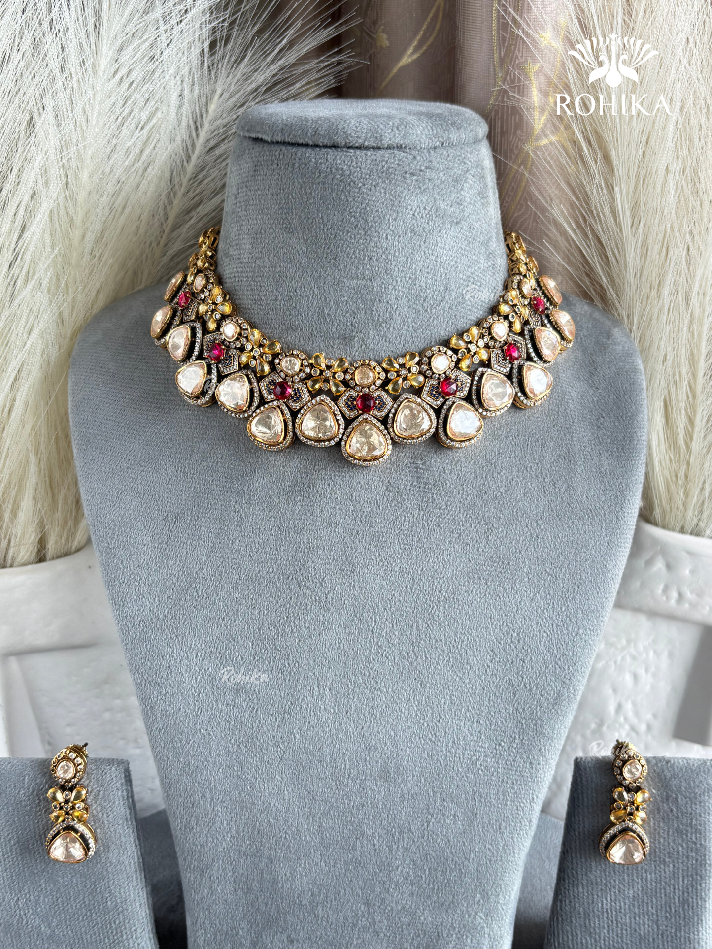 A gold-toned traditional Indian statement necklace with matching drop earrings, displayed on a gray velvet jewelry bust. The necklace features cream-colored stones, red stones, and gold-toned metal details.