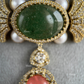 An ornate gold-toned jewelry piece featuring a large oval green cabochon stone, surrounded by small clear stones and white faux pearls, with a dangling pendant below featuring a pinkish-red oval stone.