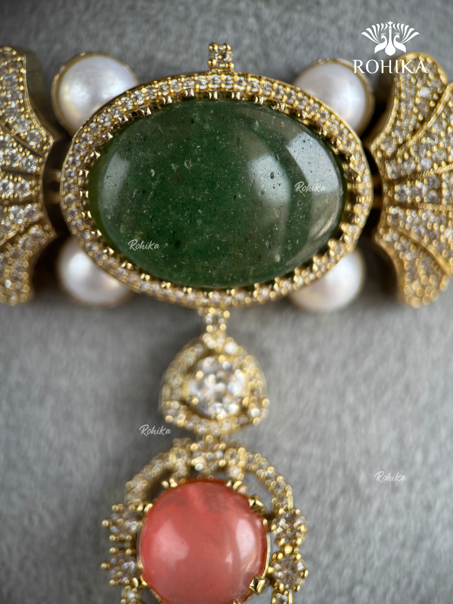 An ornate gold-toned jewelry piece featuring a large oval green cabochon stone, surrounded by small clear stones and white faux pearls, with a dangling pendant below featuring a pinkish-red oval stone.