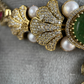 Close-up of a gold-toned jewelry piece featuring pavé-set clear stones, white pearls, and a green stone, displayed on a textured gray surface. The brand name ROHIKA is visible in the top right corner.