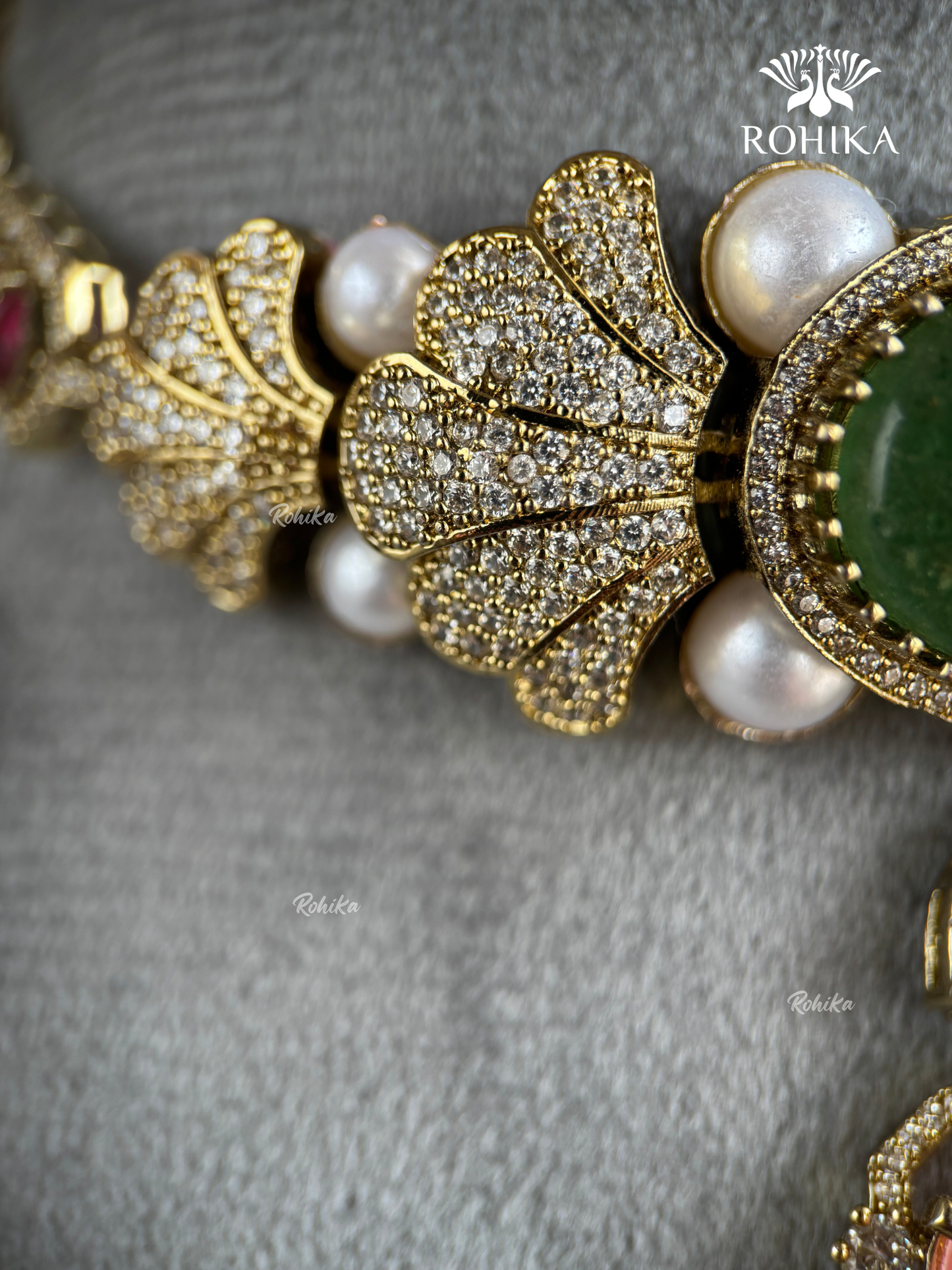 Close-up of a gold-toned jewelry piece featuring pavé-set clear stones, white pearls, and a green stone, displayed on a textured gray surface. The brand name ROHIKA is visible in the top right corner.