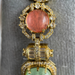A close-up of a gold-toned statement jewelry piece featuring a large round coral-pink stone at the top and a larger heart-shaped light green stone at the bottom, both surrounded by clear pavé stones on a gray velvet display.