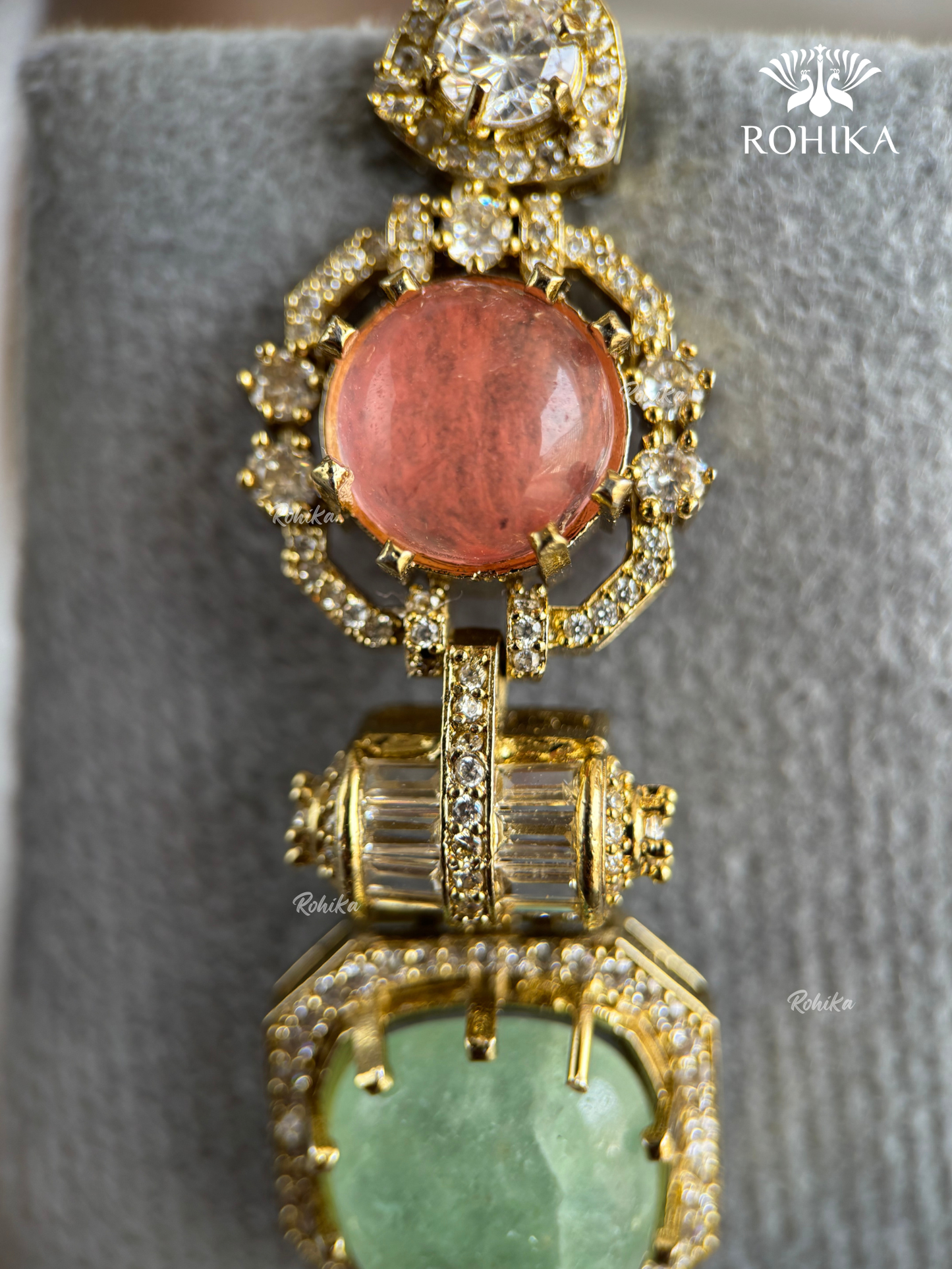 A close-up of a gold-toned statement jewelry piece featuring a large round coral-pink stone at the top and a larger heart-shaped light green stone at the bottom, both surrounded by clear pavé stones on a gray velvet display.