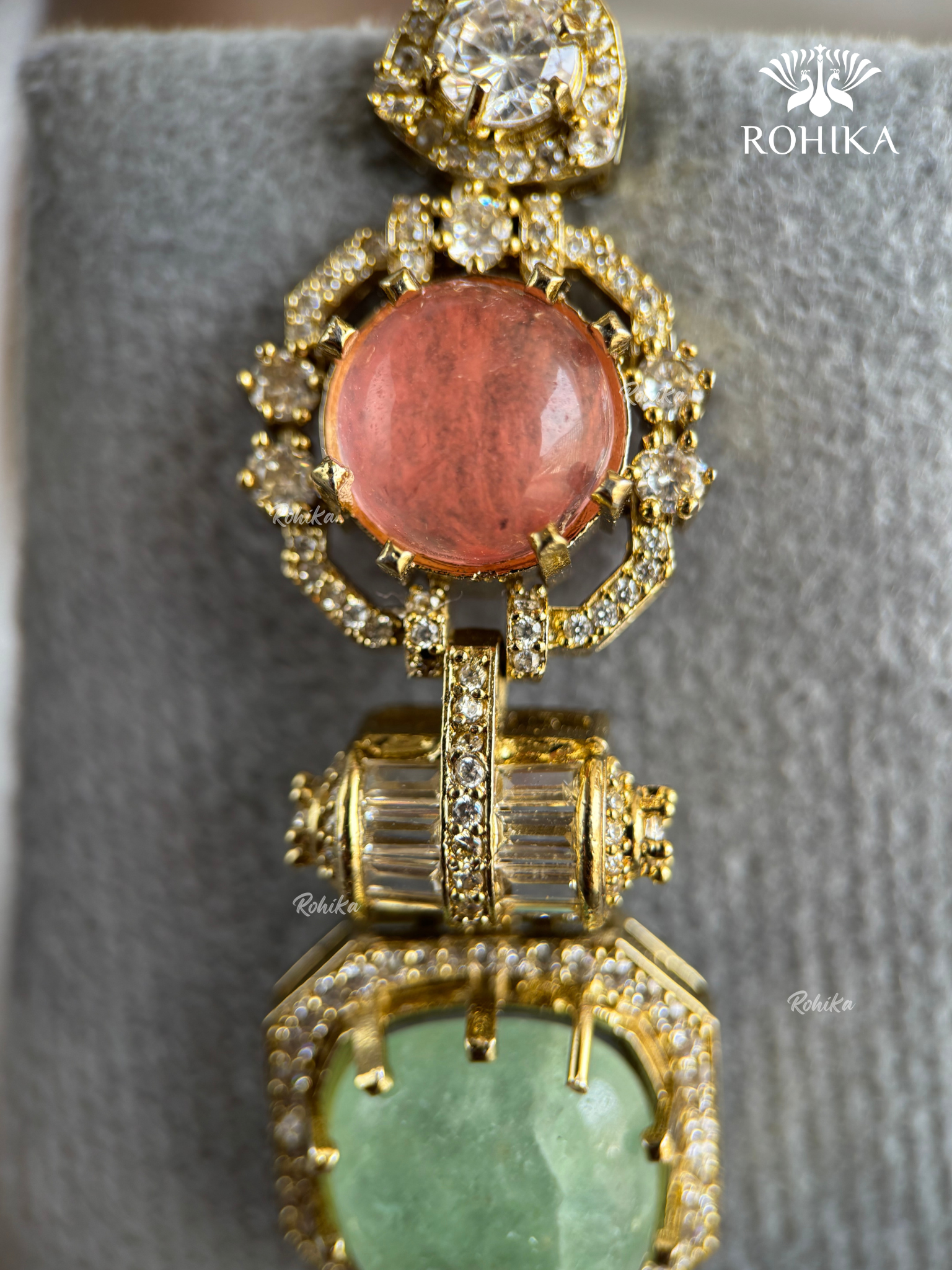 A close-up of a gold-toned statement jewelry piece featuring a large round coral-pink stone at the top and a larger heart-shaped light green stone at the bottom, both surrounded by clear pavé stones on a gray velvet display.